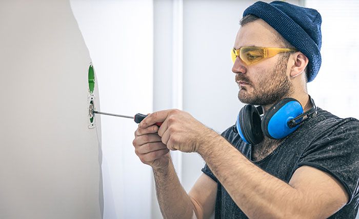 Man wearing protective glasses and ear muffs working with a screwdriver on wall wiring, representing men anonymously revealing secrets.