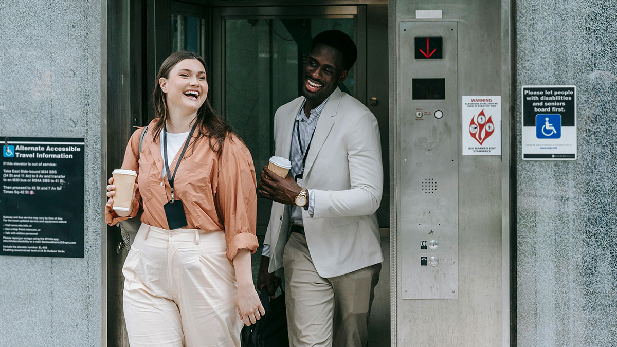 Two diverse coworkers laughing and holding coffee cups near elevator, illustrating common knowledge surprises in daily life.