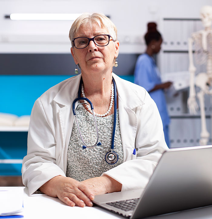 Elderly female doctor with stethoscope sitting at desk in medical office with laptop and nurse in background