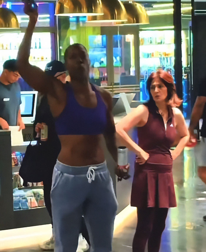 Person in athletic wear raising arm inside gym while another person watches, highlighting lesbian removed from gym controversy. Person in athletic wear raising arm inside gym while another person watches, highlighting lesbian removed from gym controversy.
