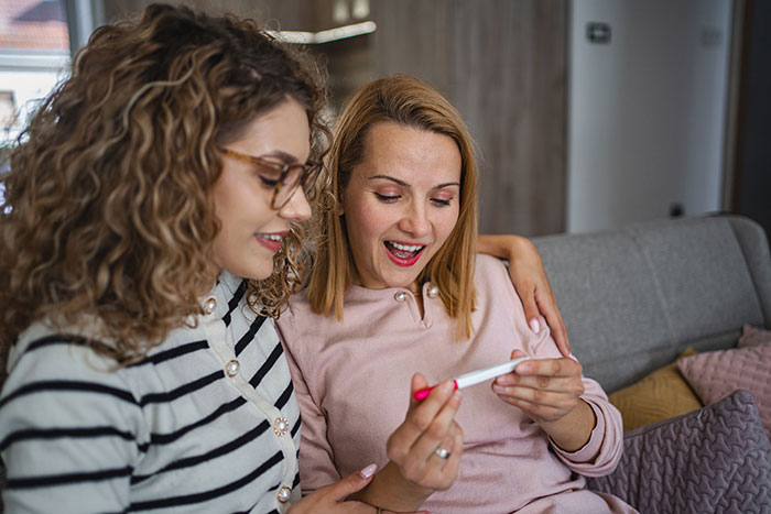 Two women on a couch, one showing a pregnancy test while the other reacts with surprise and concern.