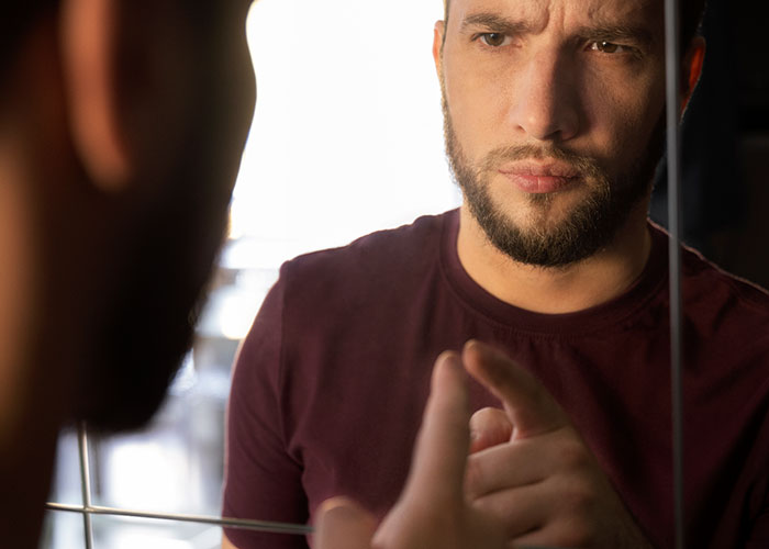 Man with beard looking intently at his reflection in the mirror, contemplating strange things neighbors accidentally saw.
