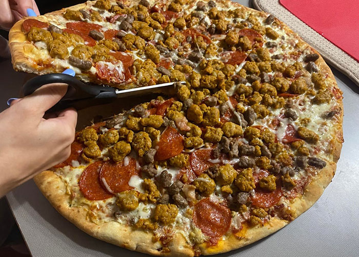 Large pizza topped with pepperoni and sausage being cut with scissors in a strange neighbor sighting scenario.