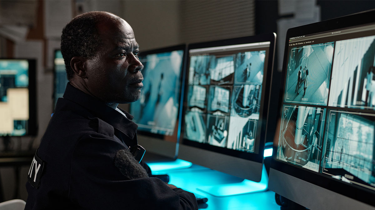 Security guard monitoring multiple surveillance camera feeds showing suspicious activity in a control room environment.