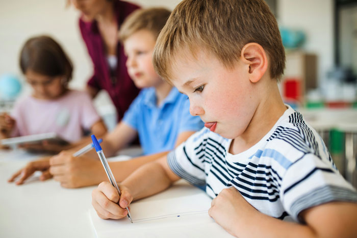Young boy focused on writing a letter with kids and teacher in the background, illustrating letters kids send to U.S. soldiers. Young boy focused on writing a letter with kids and teacher in the background, illustrating letters kids send to U.S. soldiers.