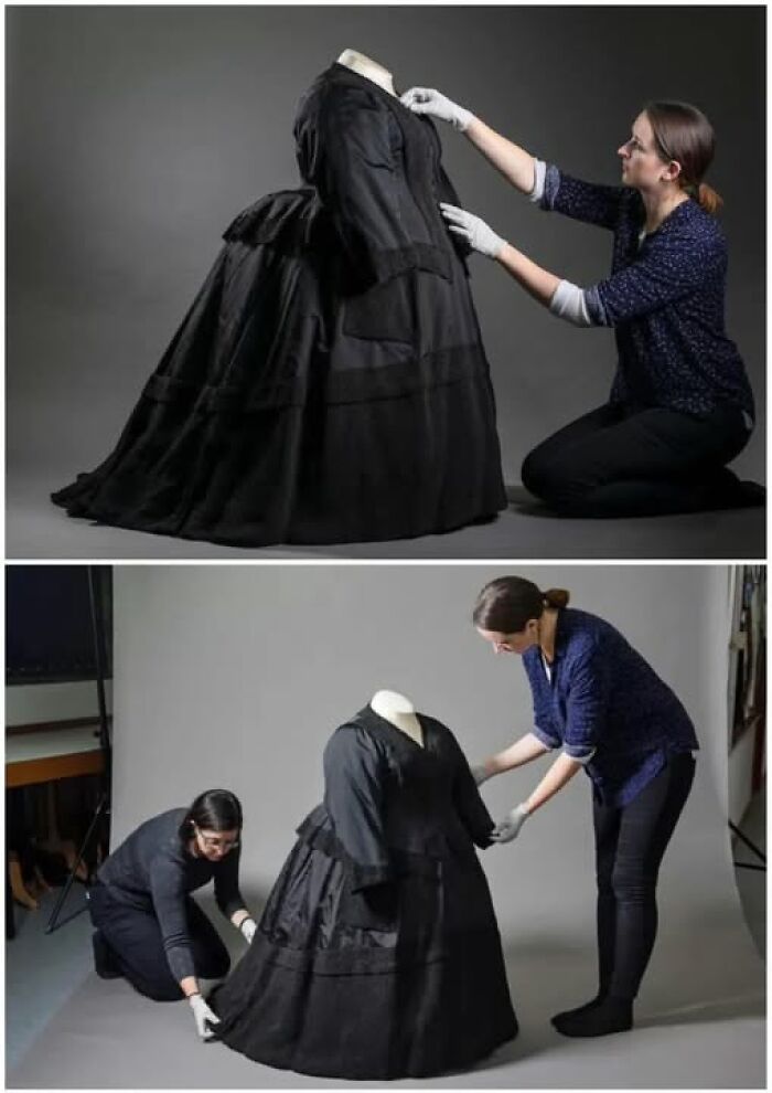 Two women carefully handling a Victorian-Edwardian dress on a mannequin in a professional studio setting.