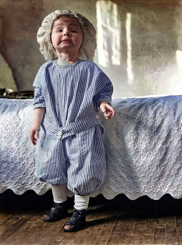 Victorian-Edwardian-Pictures of a young child dressed in striped vintage clothing and bonnet standing beside a bed in a rustic room.