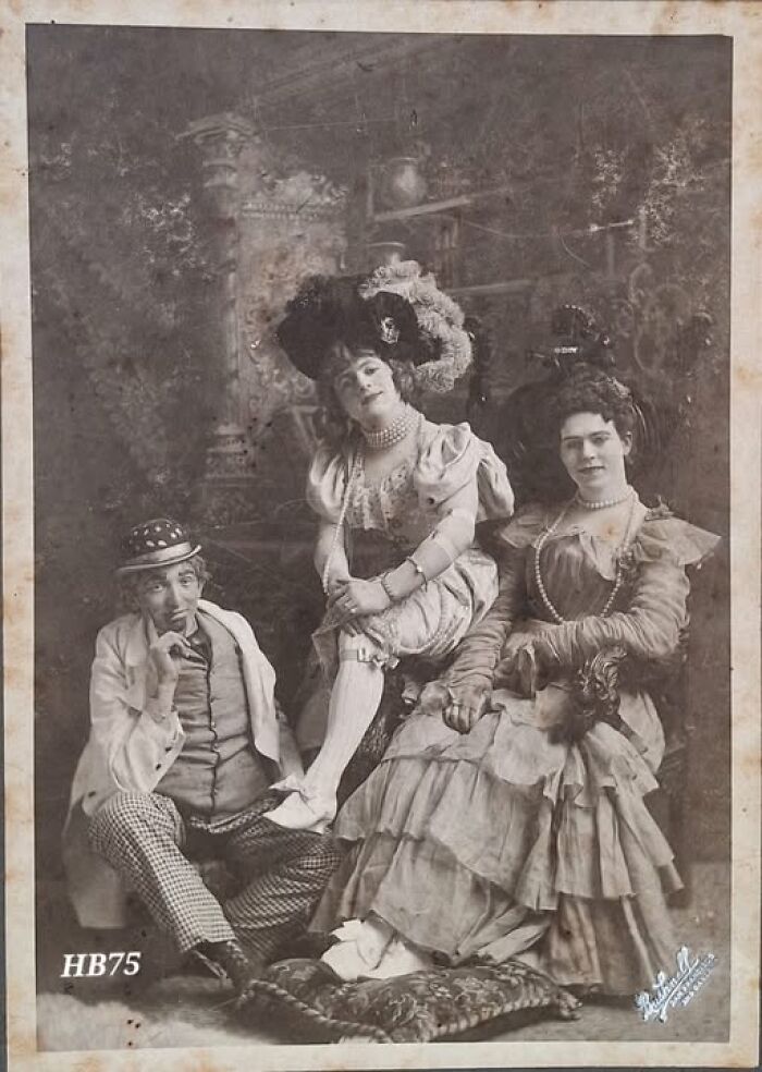 Three Victorian-Edwardian figures in elaborate period costumes and hats posing in a vintage sepia-toned photograph.