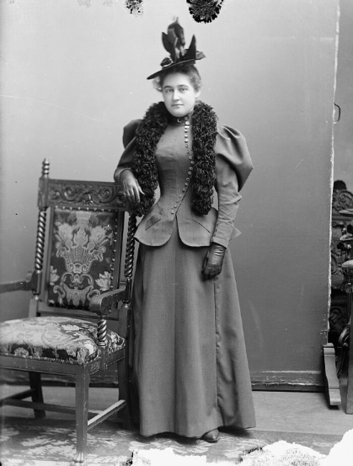 Victorian-Edwardian picture of a woman in period dress with a feathered hat standing beside an ornate chair.