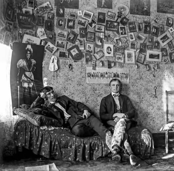 Two young men relaxing in a Victorian-Edwardian room filled with numerous vintage pictures on the wall.