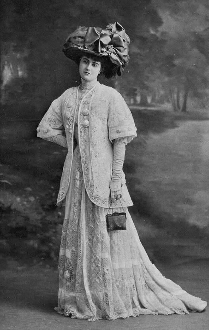 Victorian Edwardian woman in an elaborate lace dress and large decorative hat posing with small handbag outdoors.