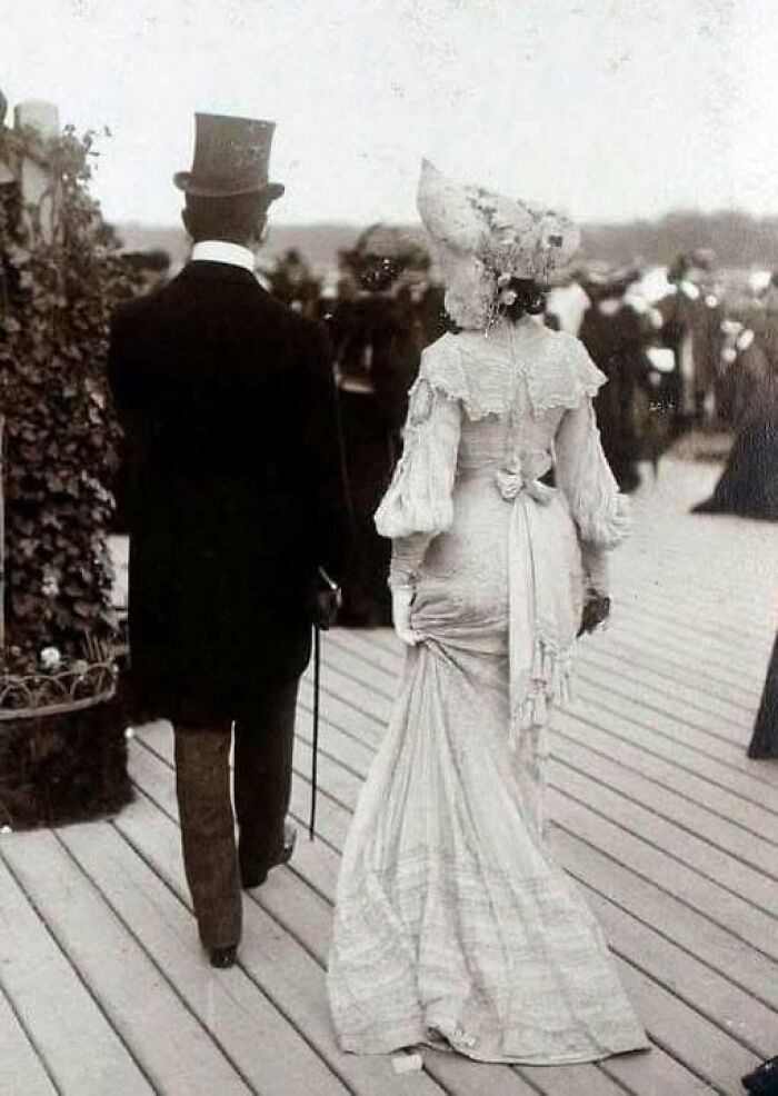 Couple dressed in Victorian Edwardian period attire walking on a wooden deck with a crowd in the background.