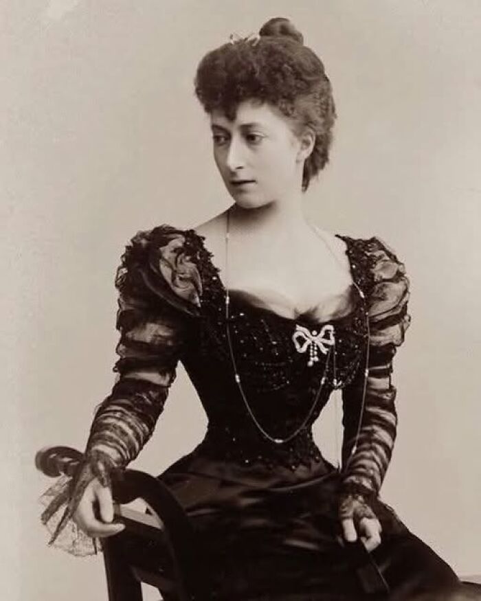 Victorian-Edwardian picture of a woman in a black vintage dress with puffed sleeves seated on a wooden chair.