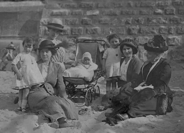 Group of women and children in Victorian Edwardian pictures sitting on the sand with a baby in a vintage pram near a stone wall.