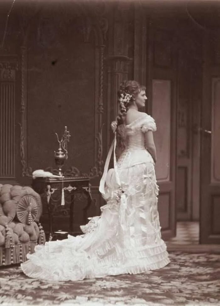 Victorian Edwardian picture of a woman in an ornate white dress with a long train standing in a vintage room.