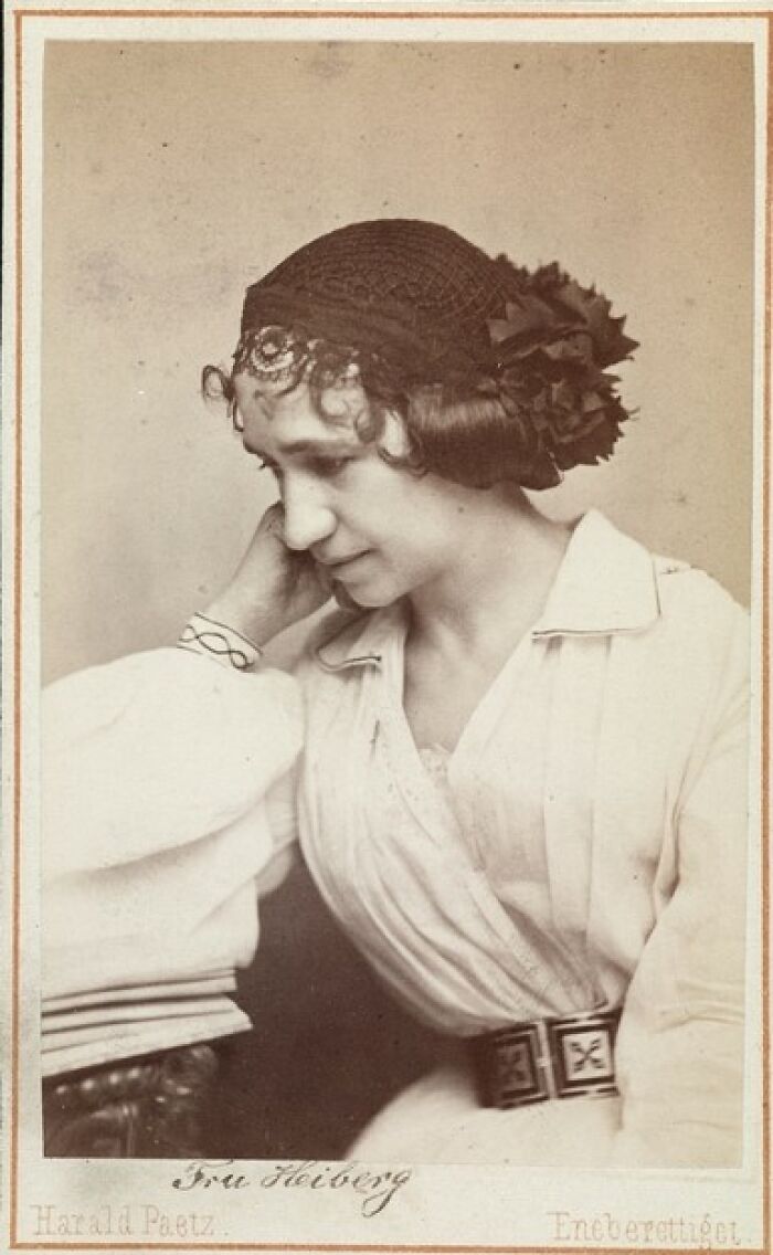 Victorian-Edwardian picture of a woman in period clothing wearing a decorated hat, resting her head on her hand.