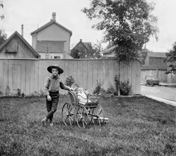 Young boy with a hat standing next to a baby in a vintage pram in a backyard, Victorian-Edwardian pictures style.