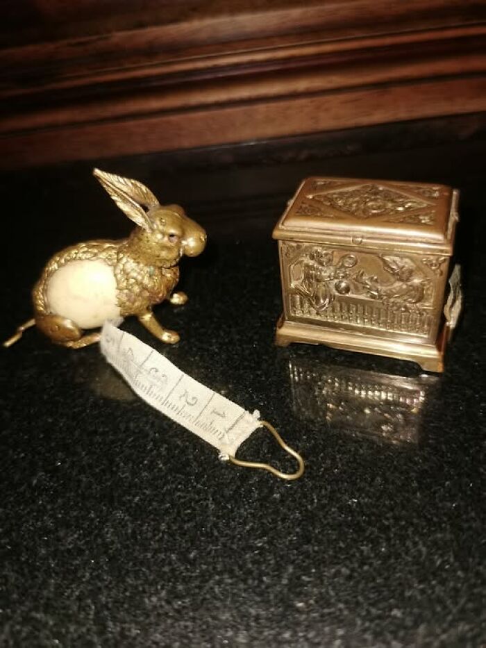 Victorian-Edwardian picture of a vintage gold rabbit tape measure next to a decorative ornate metal box on a dark surface.