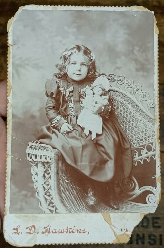 Young girl in Victorian-Edwardian style dress sitting on a wicker chair holding a doll in vintage sepia picture.