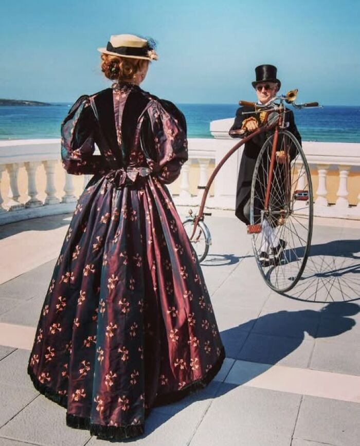 Woman in a Victorian dress and man in Edwardian attire with a vintage bicycle by the seaside in Victorian-Edwardian pictures style.