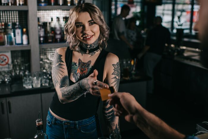 Woman with tattoos smiling and handing a card at a dimly lit bar, capturing a moment of coincidence and connection.