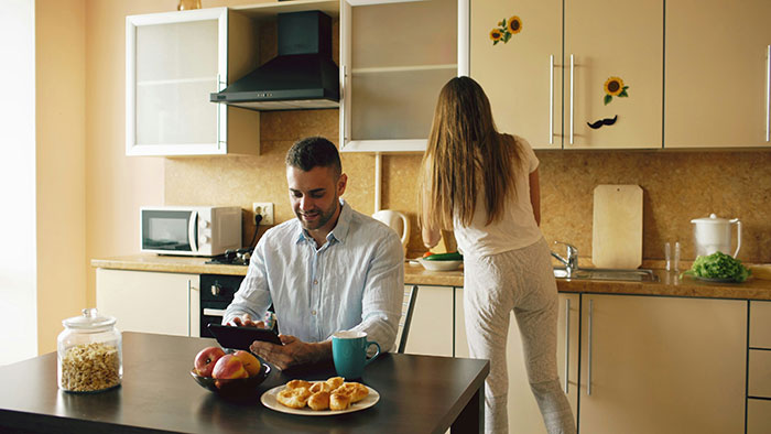 Man relaxing with tablet while woman does domestic chores in kitchen, highlighting unequal household responsibilities. Man relaxing with tablet while woman does domestic chores in kitchen, highlighting unequal household responsibilities.
