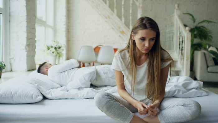 Young woman sitting on bed looking upset while her significant other sleeps, illustrating signs of unfaithful behavior.