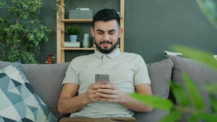 Man sitting on a couch, looking at his phone with a slight smile, hinting at subtle signs of cheating.
