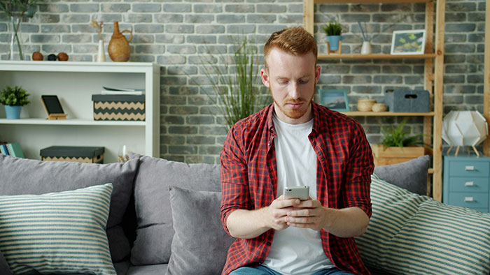 Young man in red plaid shirt sitting on couch distracted by phone, ignoring domestic chores and wife’s expectations at home. Young man in red plaid shirt sitting on couch distracted by phone, ignoring domestic chores and wife’s expectations at home.