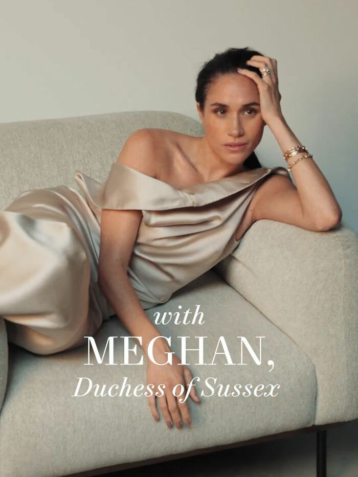 Meghan Markle posing makeup-free on a beige couch wearing a satin off-shoulder dress, looking thoughtful and relaxed.