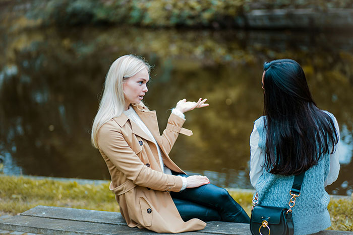 Two women sitting outdoors by water, one gesturing while discussing avid walker doing 20K steps per day. Two women sitting outdoors by water, one gesturing while discussing avid walker doing 20K steps per day.