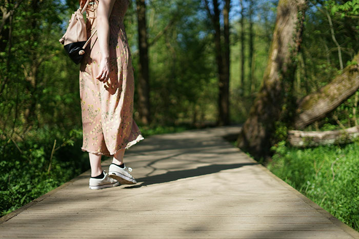 Woman walking on a wooden path in a forest wearing sneakers and a floral dress, illustrating avid walker doing 20K steps daily. Woman walking on a wooden path in a forest wearing sneakers and a floral dress, illustrating avid walker doing 20K steps daily.
