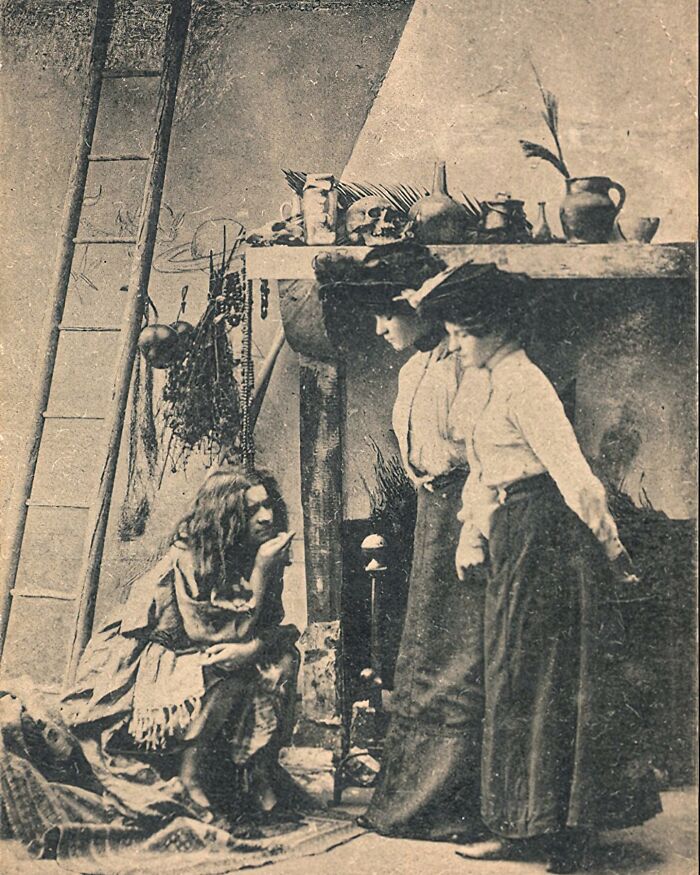 Three Victorian-era women in long skirts gathered around a seated woman in a dim, rustic interior, showcasing weird photos.
