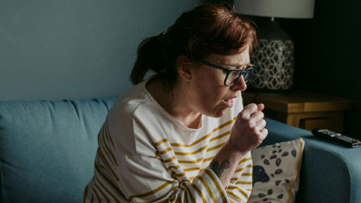 Woman wearing glasses coughing into her hand on a blue couch capturing a bizarre moment that makes people wonder sanity.