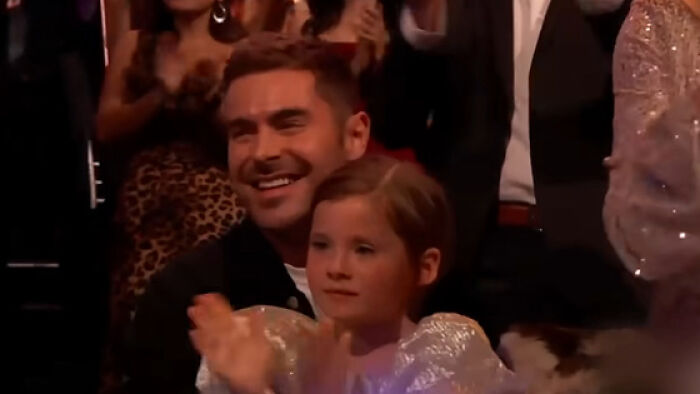 Zac Efron smiling and clapping with a child at an event, drawing attention to his back to normal face appearance. Zac Efron smiling and clapping with a child at an event, drawing attention to his back to normal face appearance.