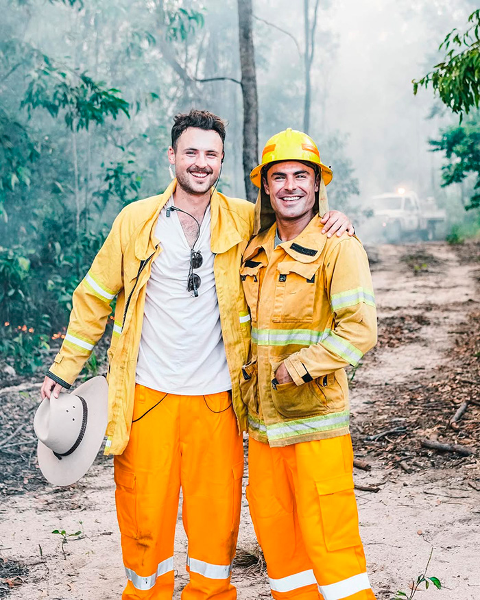 Two men in yellow firefighter gear smiling outdoors in a smoky forest related to Zac Efron's back to normal face discussion. Two men in yellow firefighter gear smiling outdoors in a smoky forest related to Zac Efron's back to normal face discussion.