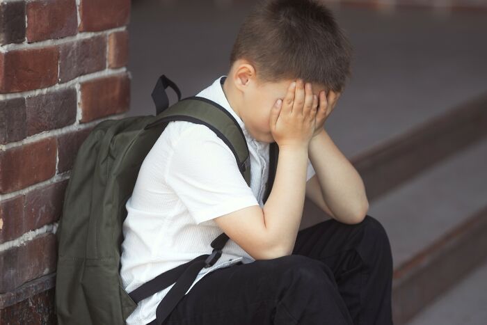 Young boy with backpack sitting on steps, covering face, illustrating emotional struggle and insensitive remarks about cancer.