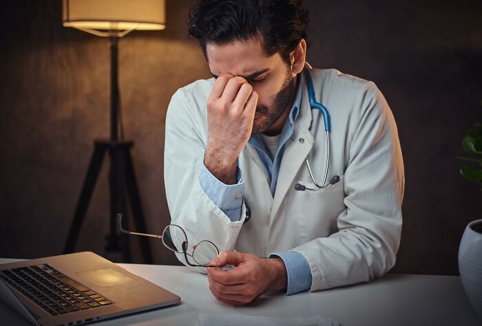 Male doctor in white coat sitting at desk, holding glasses and rubbing his eyes, showing stress and fatigue related to cancer challenges.