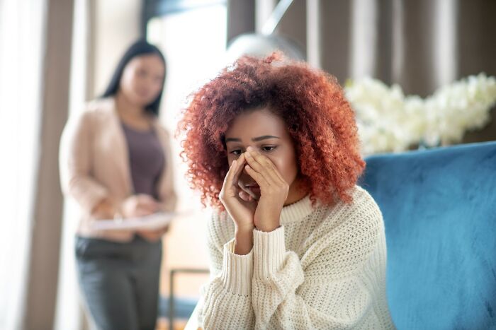 Young woman with red curly hair looking distressed while sharing cancer struggles, with a supportive person in the background.