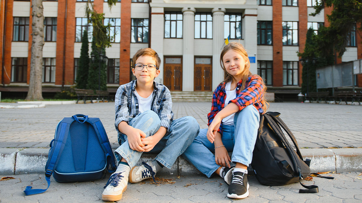 Two children sitting outside a school building with backpacks, representing stepchildren at home and boarding school.