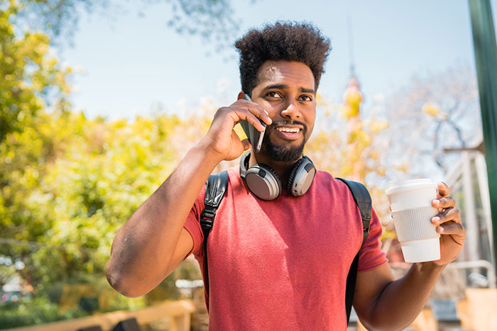 Man wearing headphones and red shirt, holding coffee and talking on phone during suspicious work trip outdoors. Man wearing headphones and red shirt, holding coffee and talking on phone during suspicious work trip outdoors.