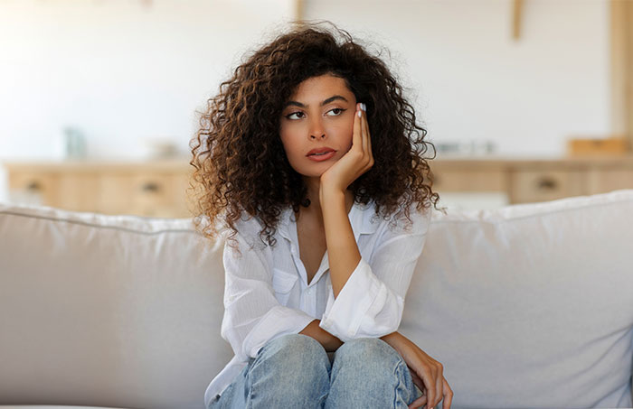 Young woman sitting on couch looking upset and thoughtful about guy secretly giving his sister a key to GF’s house