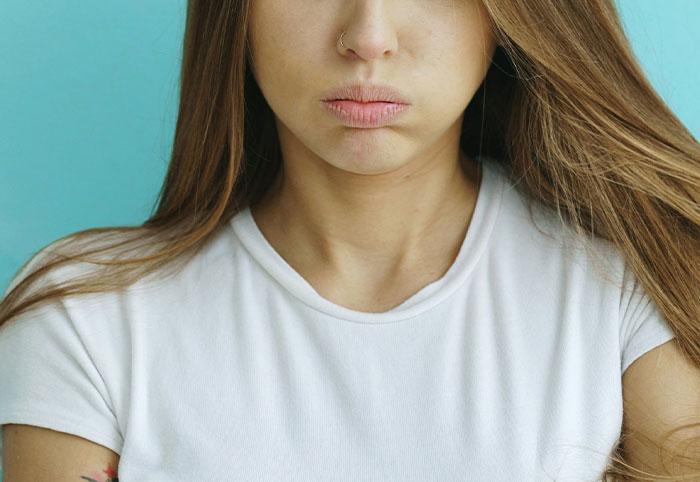 Young woman with long hair in a white shirt looking pouty against a blue background, expressing woman child emotions.