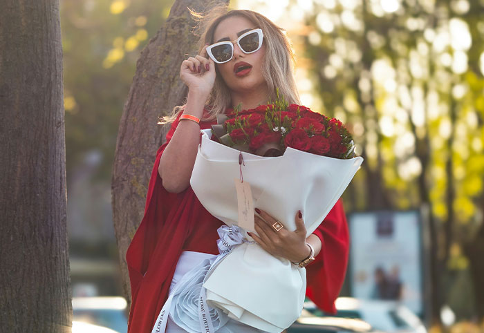 Woman child holding bouquet of red roses outdoors, wearing white sunglasses and a red outfit near a tree.
