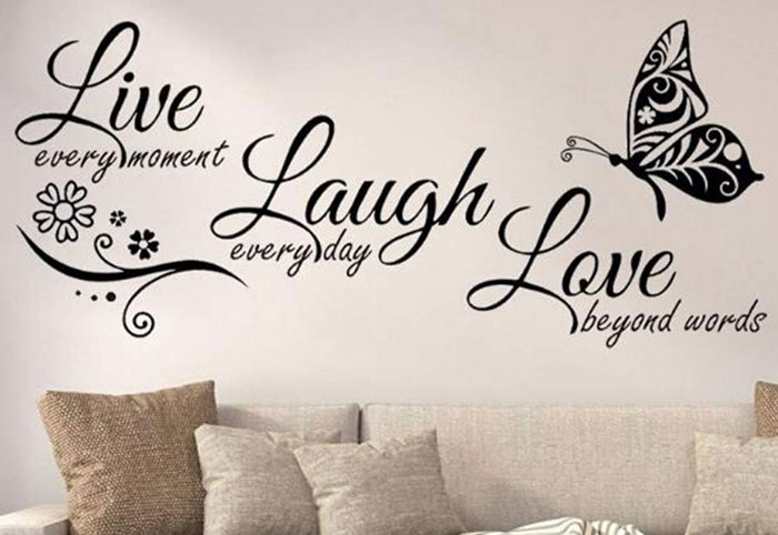 Decorative wall decal with calligraphy words Live Laugh Love and butterfly above a beige couch with cushions, woman child theme.