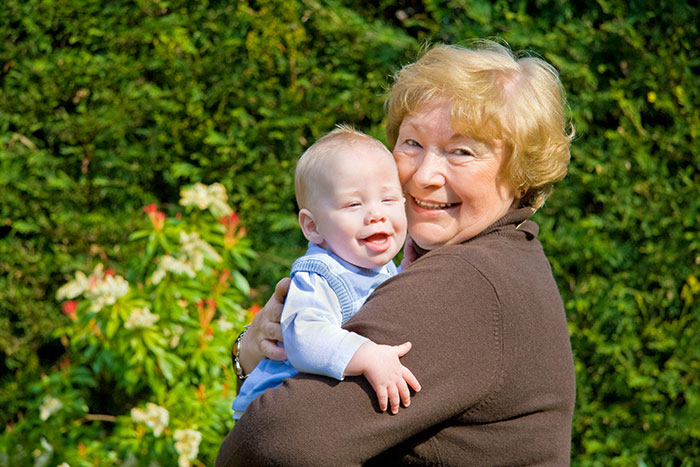 Elderly woman holding smiling baby outside, illustrating MIL admits to having a favorite grandchild and family tension. Elderly woman holding smiling baby outside, illustrating MIL admits to having a favorite grandchild and family tension.