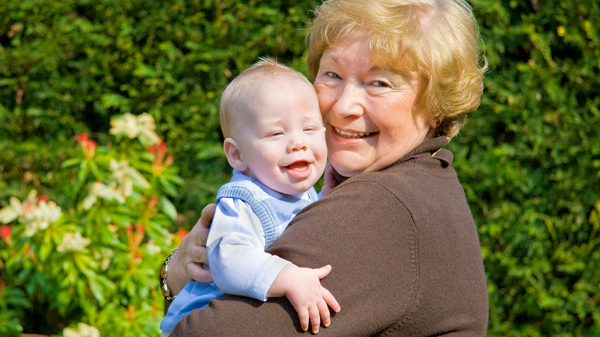 Older woman happily holding her favorite grandchild outdoors with greenery and flowers in the background.