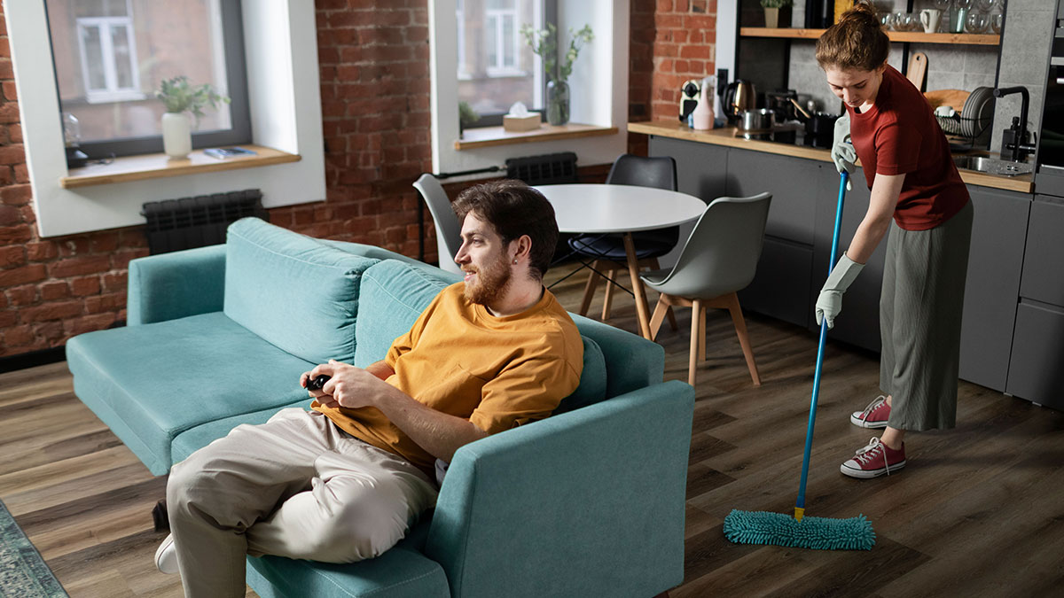 Woman mopping floor in kitchen while man lounges on sofa, illustrating jobless partner costing more than he brings in.