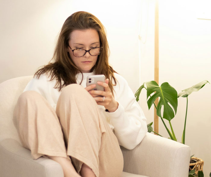 Woman in glasses sitting thoughtfully on chair, focusing on her phone, reflecting on traditional family Thanksgiving hosting. Woman in glasses sitting thoughtfully on chair, focusing on her phone, reflecting on traditional family Thanksgiving hosting.