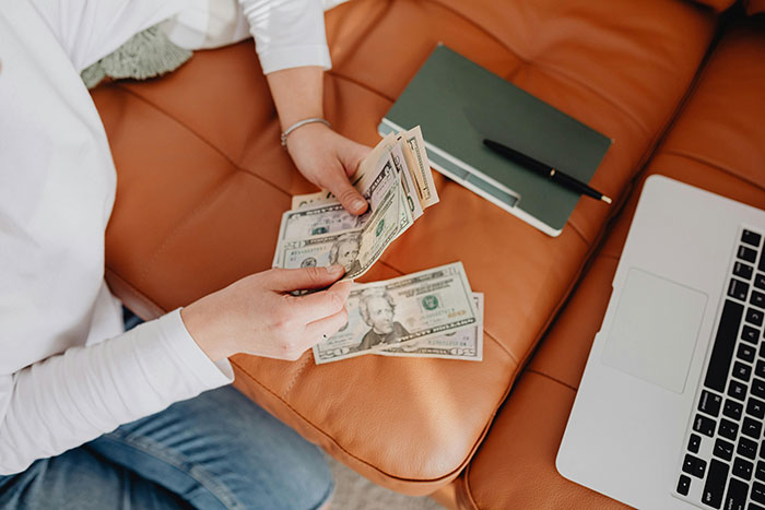 Woman counting cash on a couch next to a laptop and notebook, considering a $4k plastic surgery request. Woman counting cash on a couch next to a laptop and notebook, considering a $4k plastic surgery request.